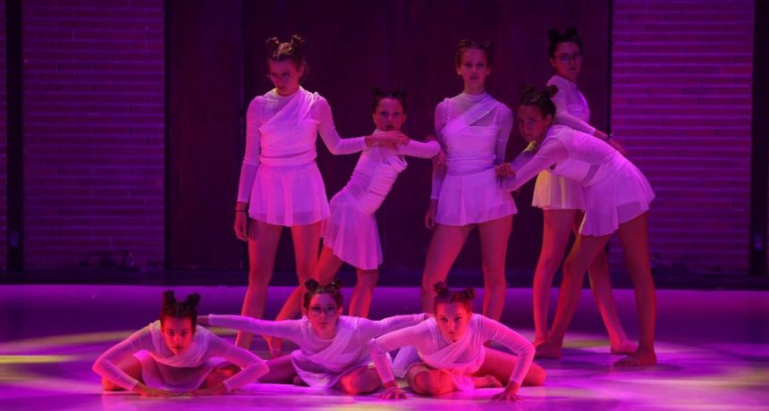 Groupe de jeunes danseurs en costume blanc sur scène colorée violette.
